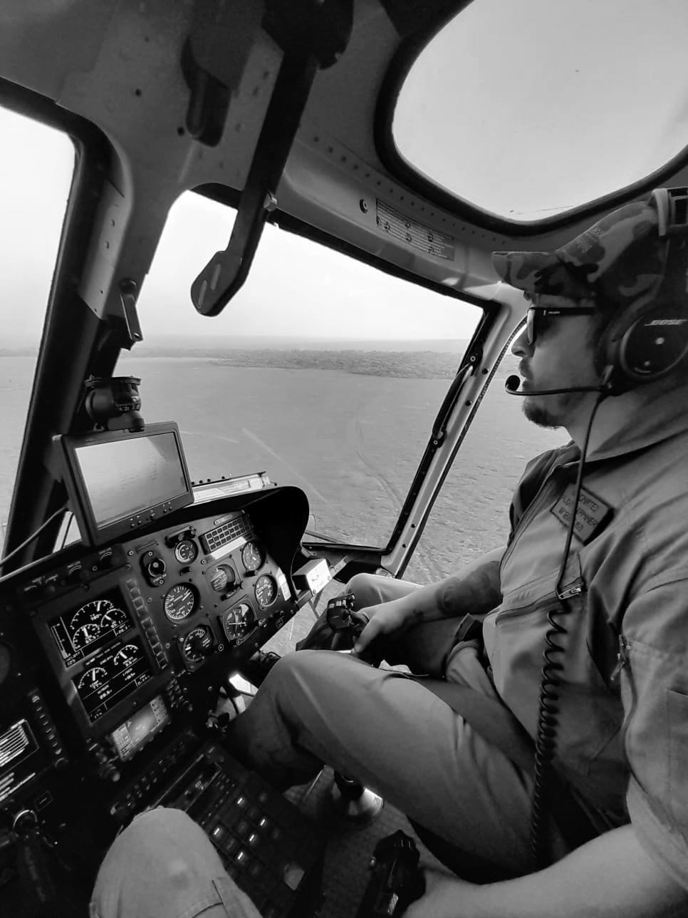 Ivan van Rooyen flying a helicopter low over an open grass plain, in a flight suit and headset.