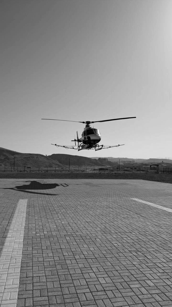 A helicopter fitted with spray booms hovering low over a paved pad, its shadow on the ground beneath it.