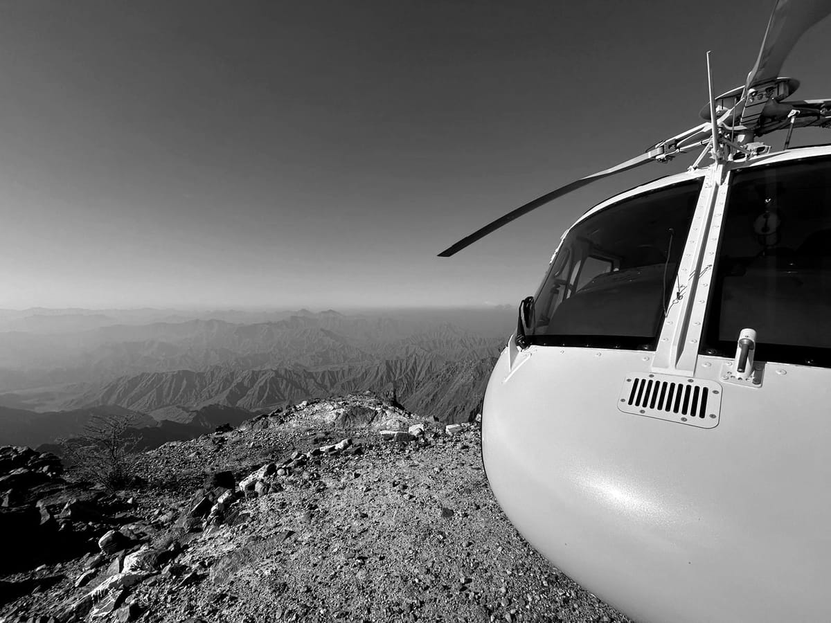 The nose and rotor head of a light helicopter parked on a rocky summit, looking out over layered mountain ridges fading into haze.
