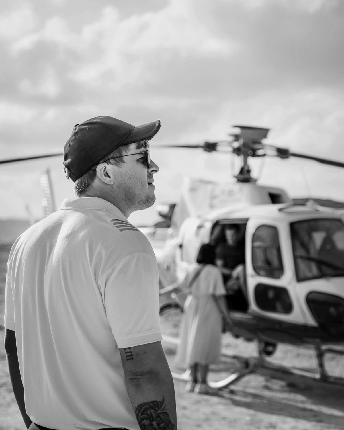 Ivan van Rooyen in a pilot shirt, cap and sunglasses, seen in profile as passengers board a helicopter behind him.