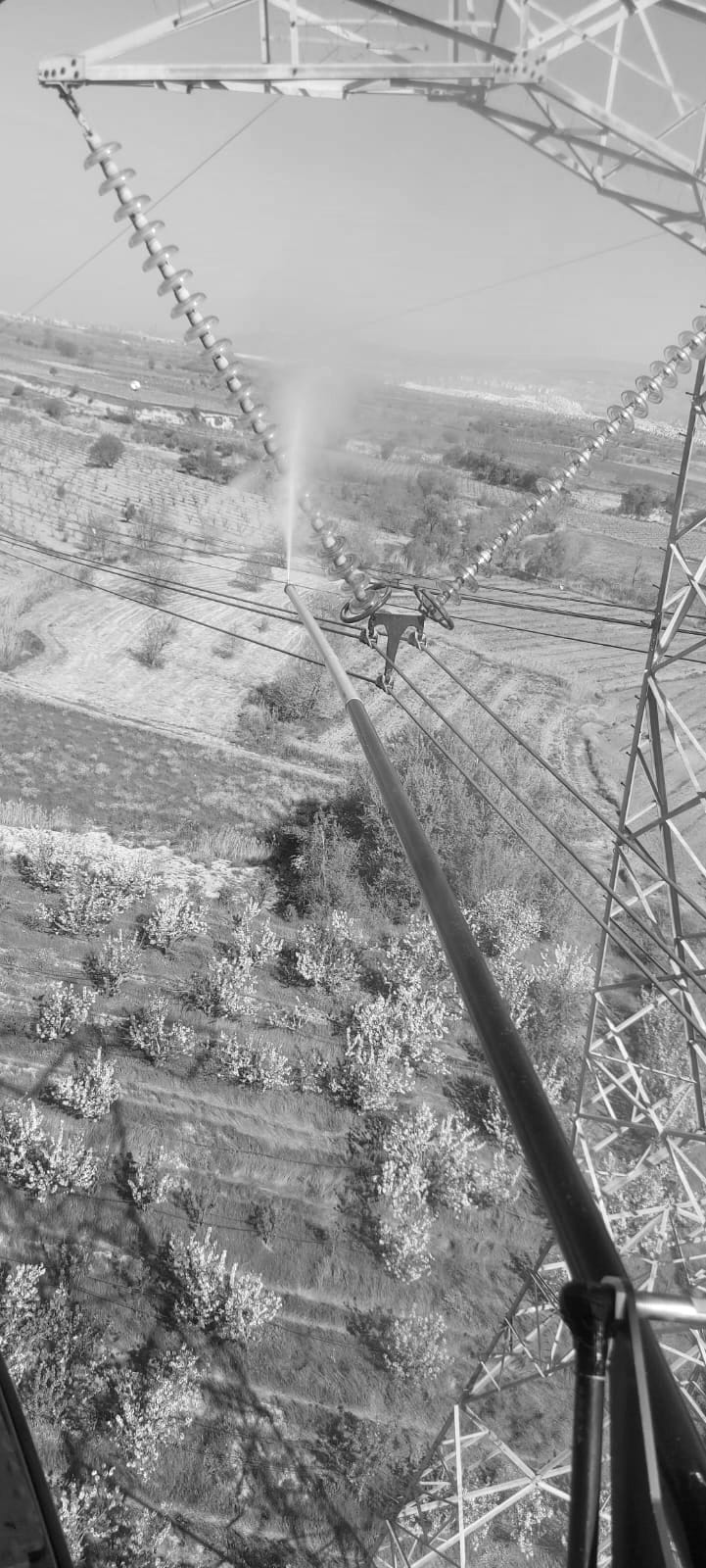 View from a helicopter of a wash lance spraying water onto a transmission-line insulator string, with orchards far below.