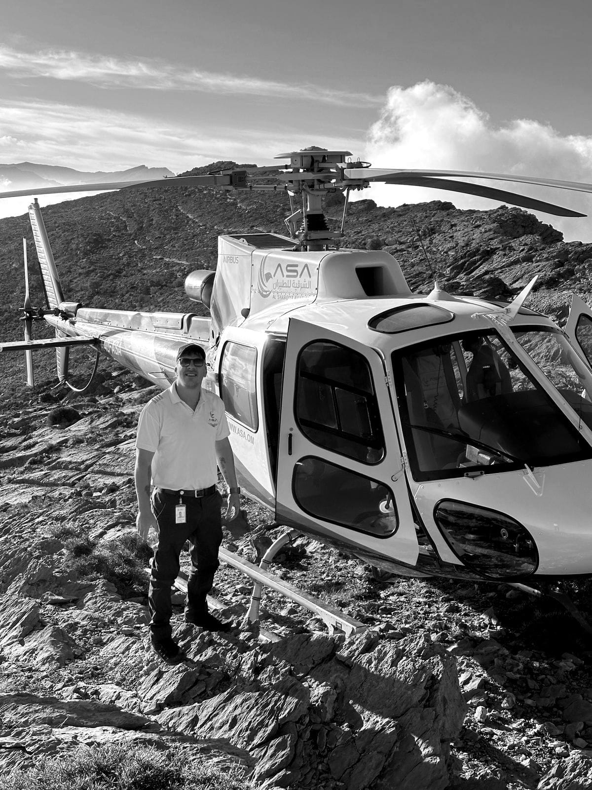 Ivan van Rooyen standing beside a helicopter parked on a rocky mountain ridge, with cloud below the ridgeline.