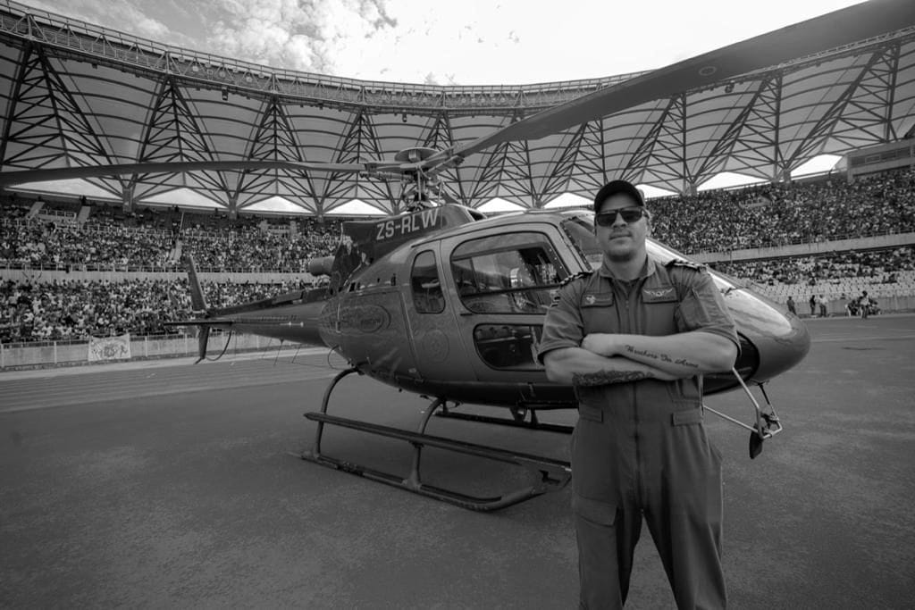 Ivan van Rooyen in a flight suit, arms folded, in front of a helicopter parked on the running track of a full stadium.