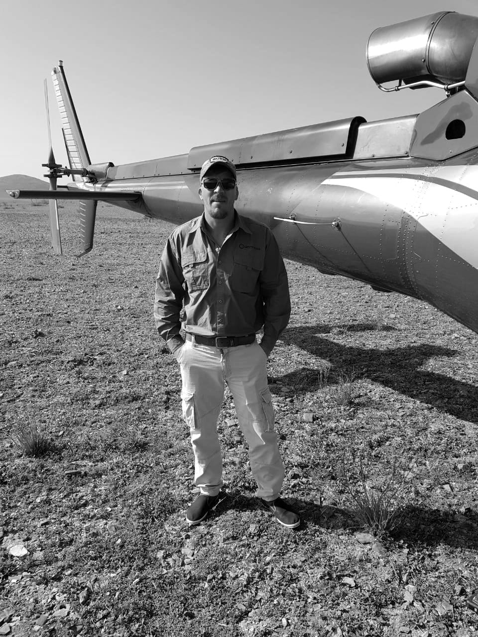 Ivan van Rooyen standing in open veld beside the tail boom of a helicopter.